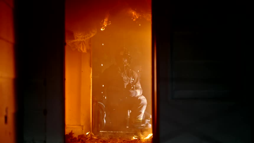 A firefighter with a green laser pointer on the helmet sits amidst orange flames and smoke inside a structure as seen from a doorway