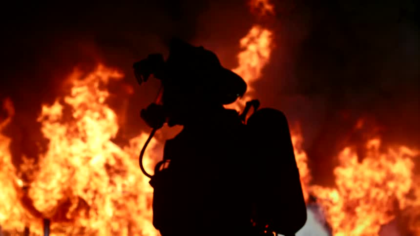 A firefighter in full gear including a breathing apparatus stands silhouetted against a backdrop of intense flames engulfing a building