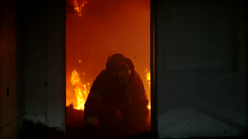 Firefighter crouches forward through a door as flames consume the interior of the room