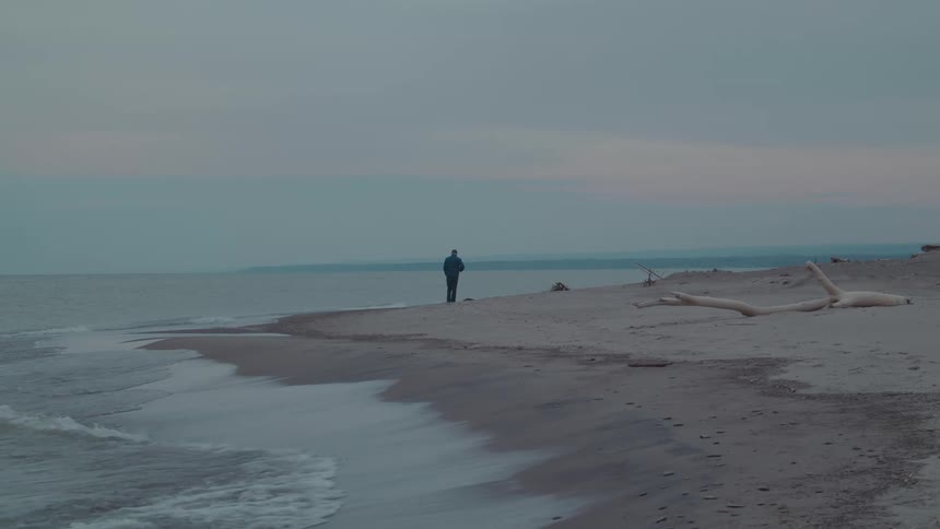 A person walks alone on a quiet beach gazing out at the calm ocean under a cloudy sky