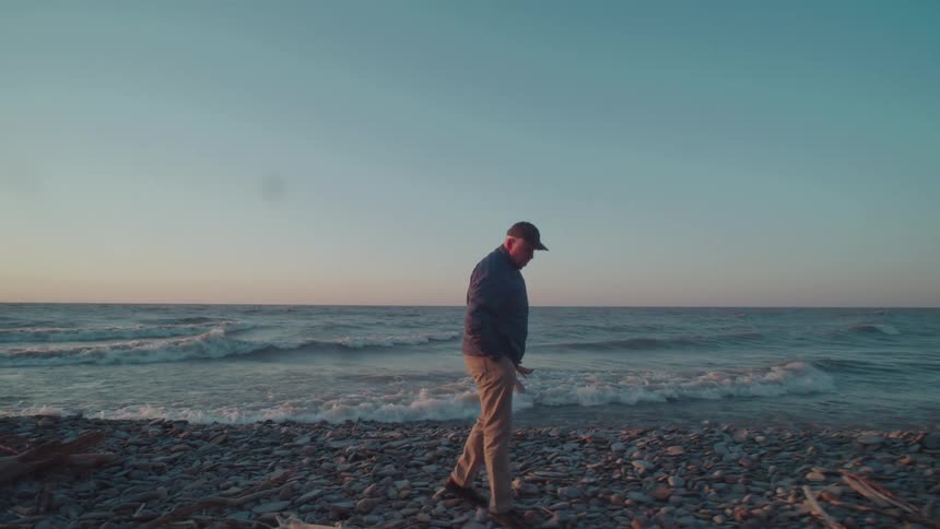 An elderly man wearing a hat and jacket walks on a rocky beach as ocean waves hit the shore