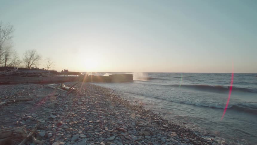 Moving across a rocky beach covered in pebbles and driftwood as gentle waves lap the shore during a bright sunrise