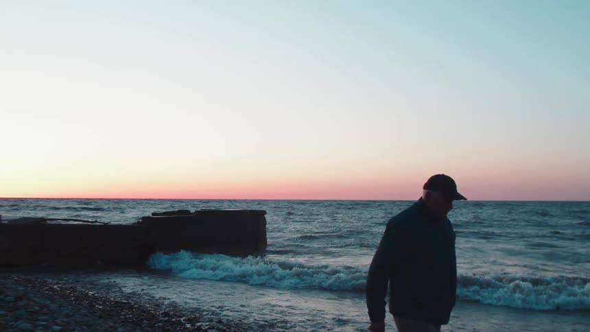 A man walks along the rocky shore at sunset as waves crash beside him