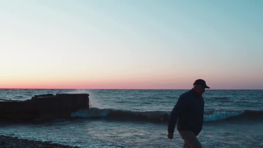 A man wearing a hat and jacket walks on the beach at sunrise as ocean waves hit the shore
