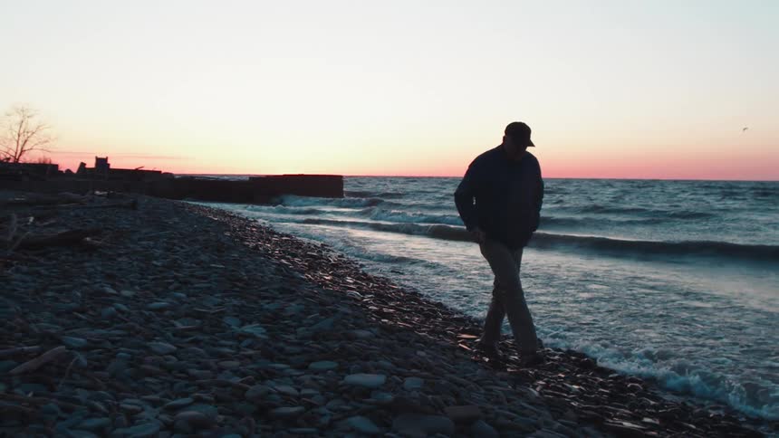 A man walks alone along a rocky beach at sunset with waves gently crashing nearby