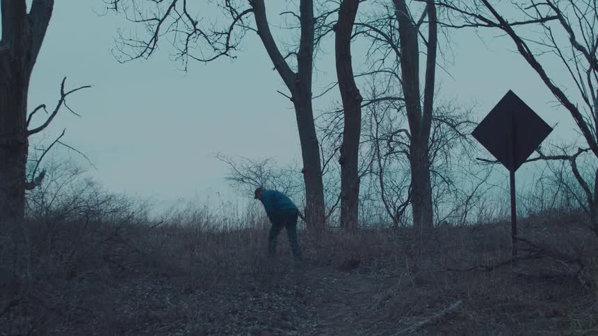 A man wearing a blue jacket and a hat hikes through the woods walking past a signboard