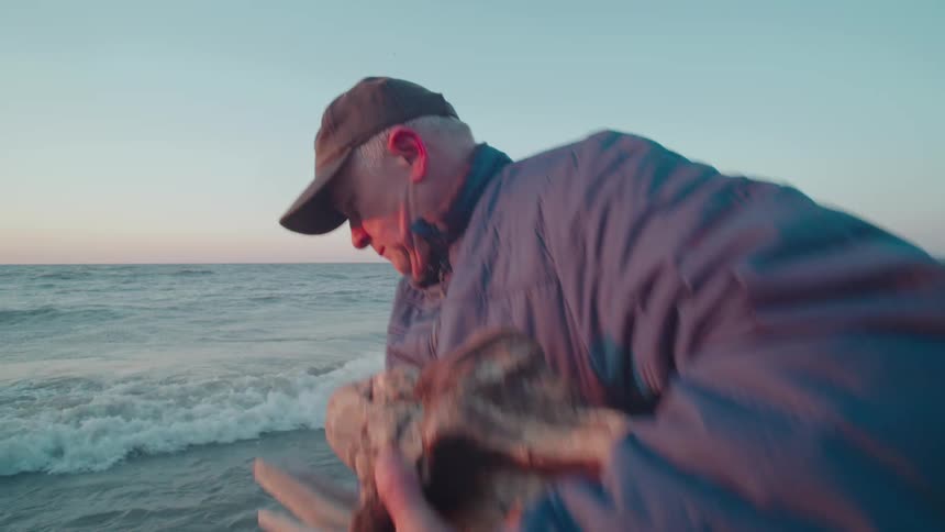 Man in cap carrying pieces of tree trunk walking along seashore