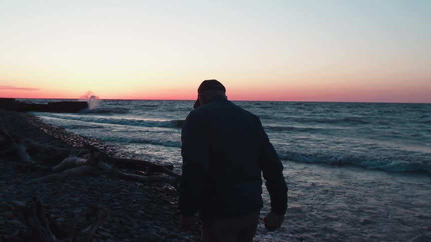 A man walks alone along a rocky beach at sunset facing the ocean waves