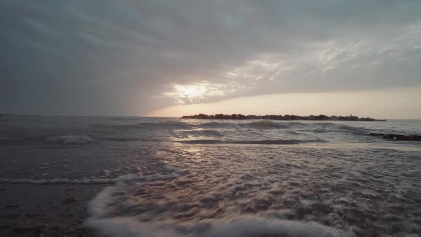 Sunset light reflects off gentle waves as they wash over a rocky shoreline under a cloudy sky