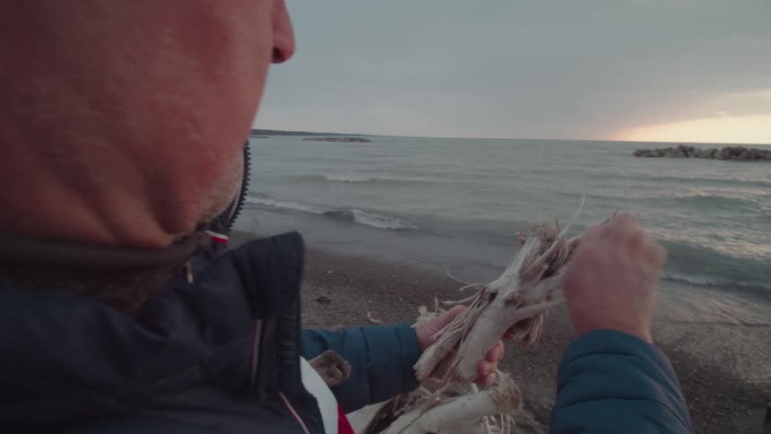 Man on a beach peeling a tree branch