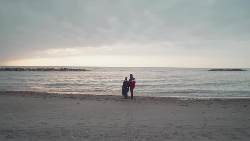 Man and woman stand together on the beach as he holds a piece of driftwood pointing out to the water as waves roll in to shore