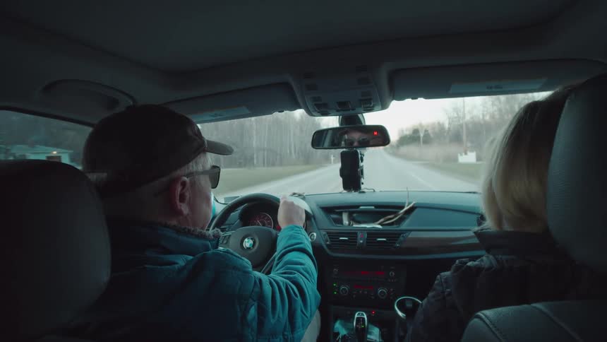Elderly man converses with a woman in a car as he drives down a road during daytime