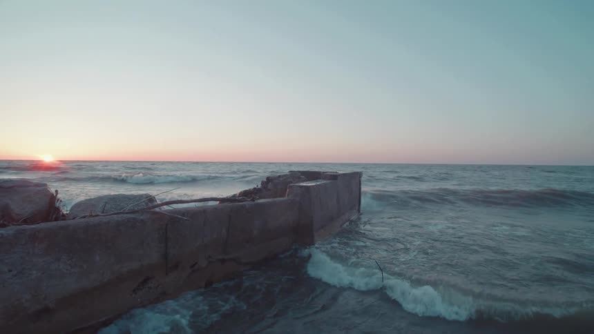 Ocean waves crash against a worn concrete pier at sunset