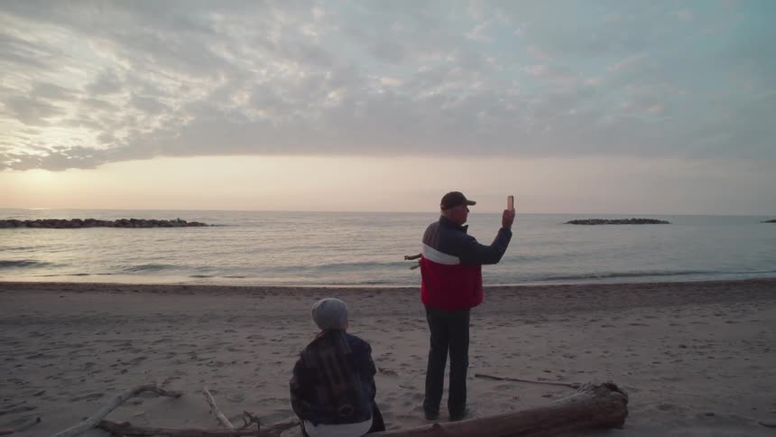 Two people on the beach at sunrise with a man standing filming with a smartphone