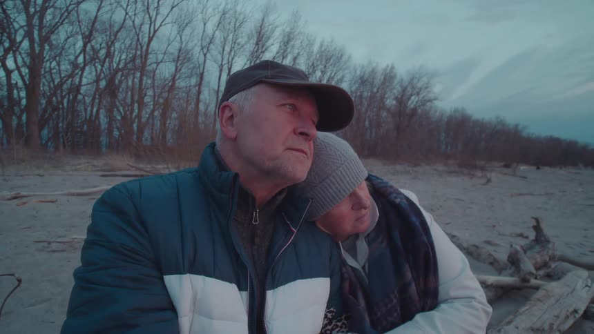 An older couple sits closely together on a quiet sandy beach at dusk