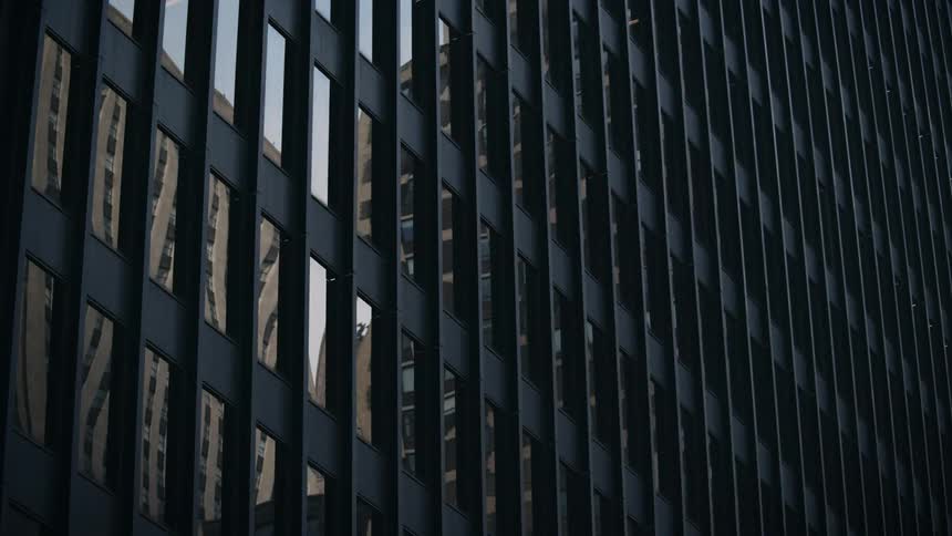 Sunlight passes over buildings in the reflections of the dark glass and steel facade of a modern skyscraper