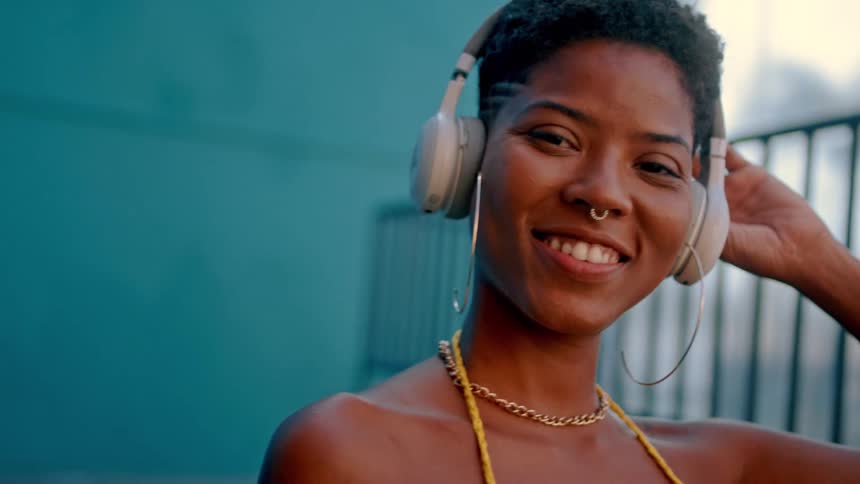A smiling woman is wearing headphones outdoors enjoying music in the sunlight