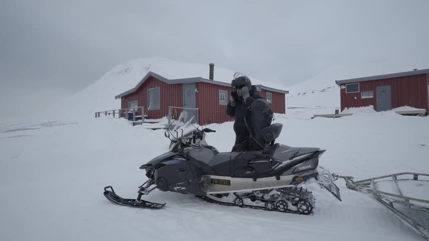 A man in heavy winter gear exits a cabin and walks toward a snowmobile