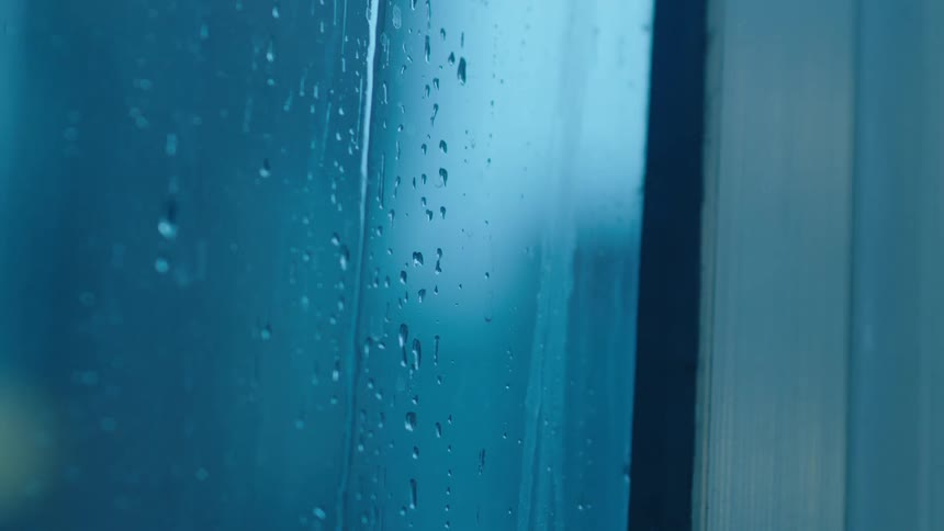 Raindrops cling to and slowly stream down a windowpane during a gloomy bluetoned day