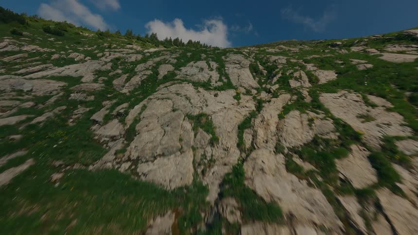 A fast paced aerial shot captures a rocky mountainside with patches of green grass and small yellow flowers under a bright blue sky with scattered white clouds