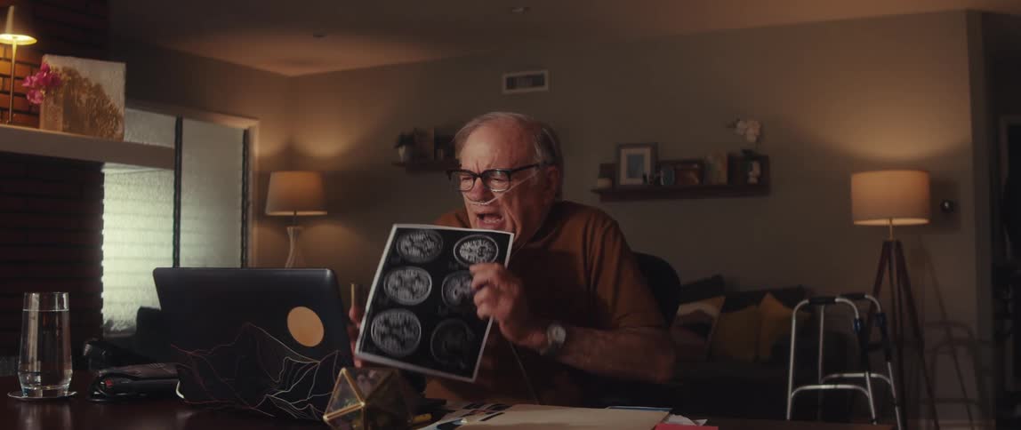 An elderly man in his cozy living room shares brain scans over a video call with a doctor on his laptop