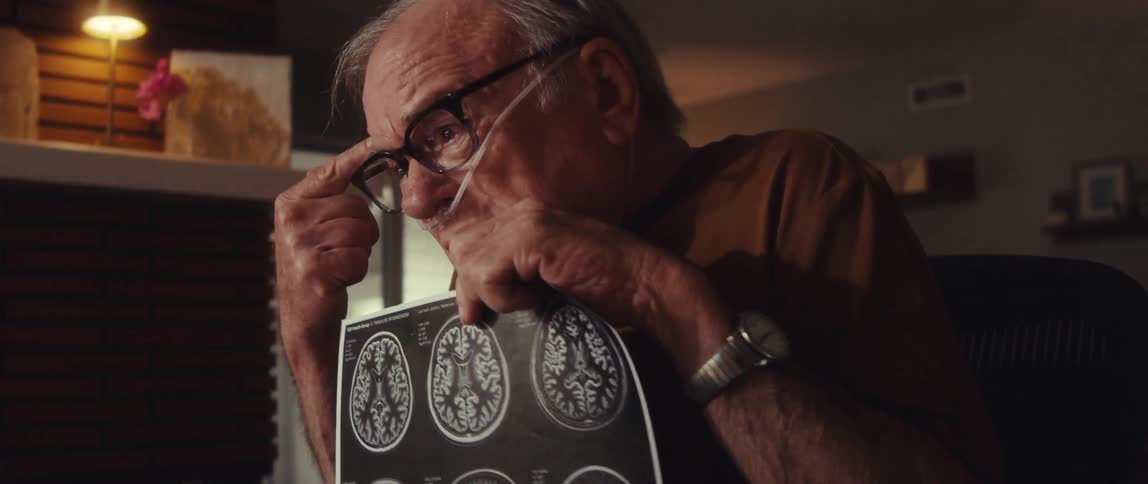An elderly man sits at a desk holding brain scan images beside a laptop appearing nervous and distressed