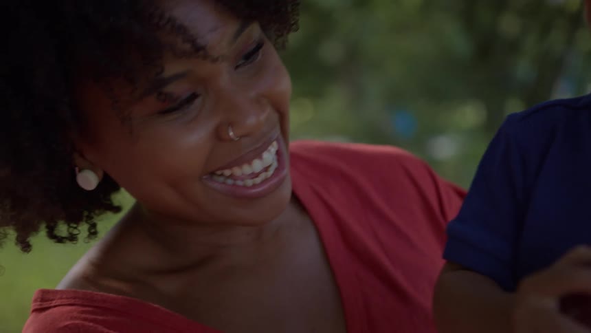 A mother in a red shirt watches as her child in blue holds a red apple outdoors amid trees and foliage