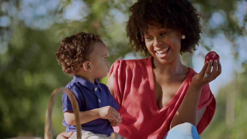 A smiling woman holds a fruit while sitting outdoors with a young child on her lap