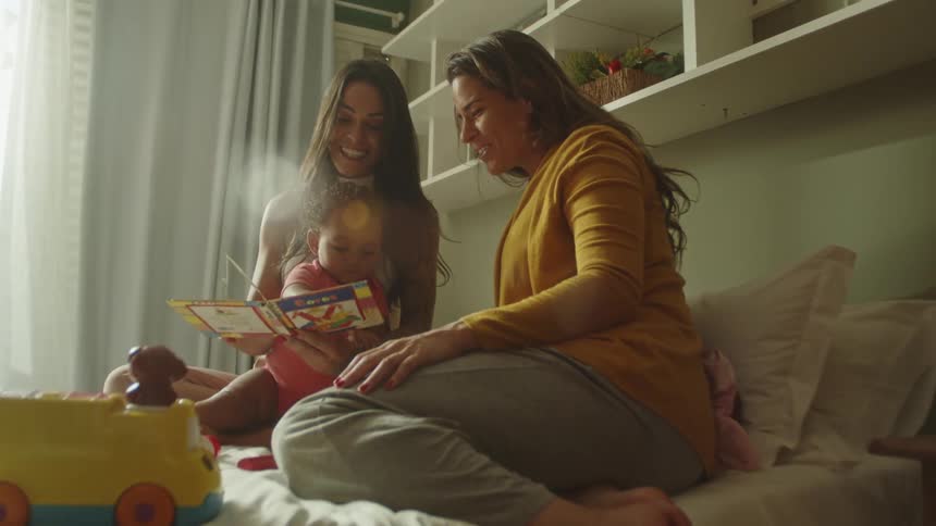 Two women and a child joyfully read a book together on a bed in a cozy room
