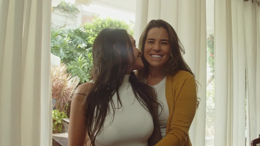 Two smiling women standing near the glass window with one kissing the other on the cheek at daytime