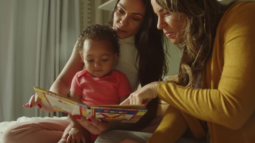 Two women sit with a baby while reading a book