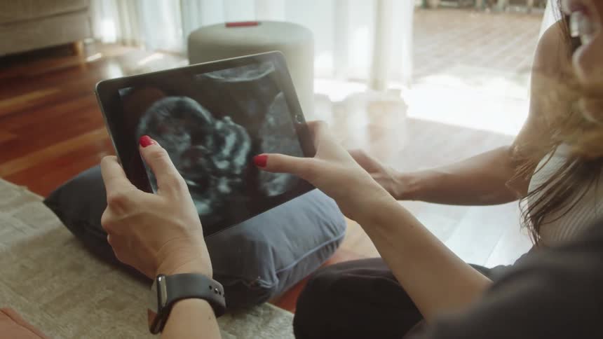 Close up of two women looking at a baby ultrasound photo on a tablet device while sitting on the floor in a room during daytime