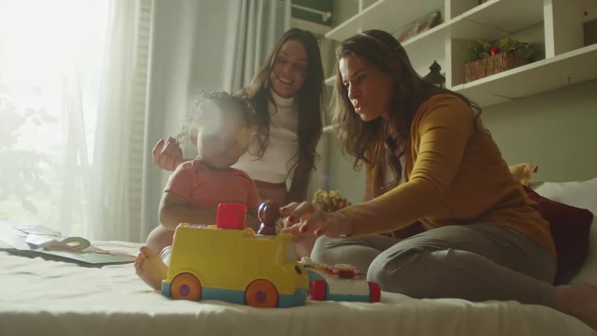 Two women play with a child who is playing with a toy all on top of the bed in the bedroom around midday