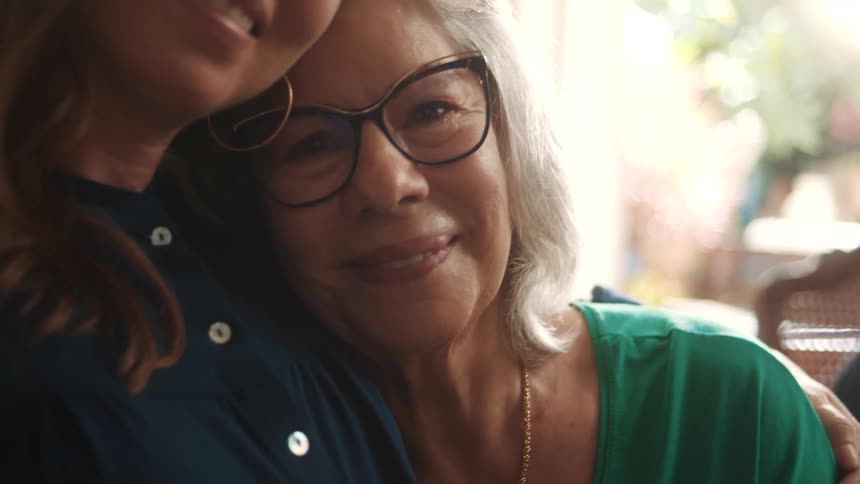 A smiling older woman wearing glasses is embraced by a person with brown hair who is partially visible