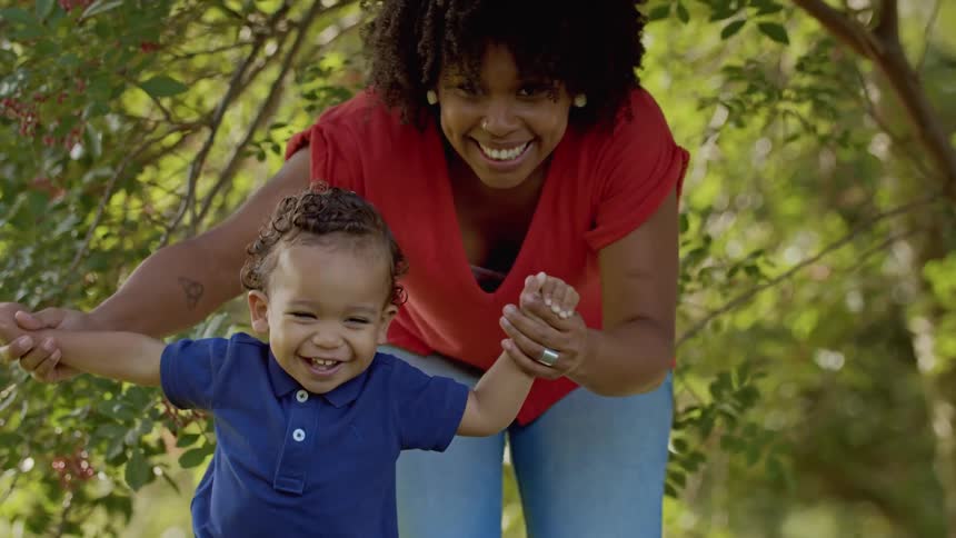 A woman joyfully helps a toddler walk through a sunlit forest path