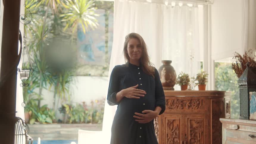 A pregnant woman stands indoors smiling and gently holding her belly