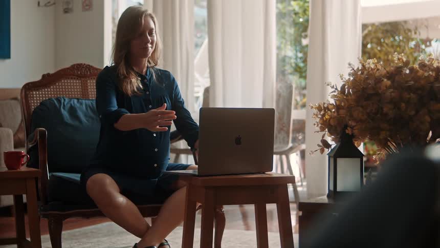 A pregnant woman sits relaxed and smiling in front of a laptop at home