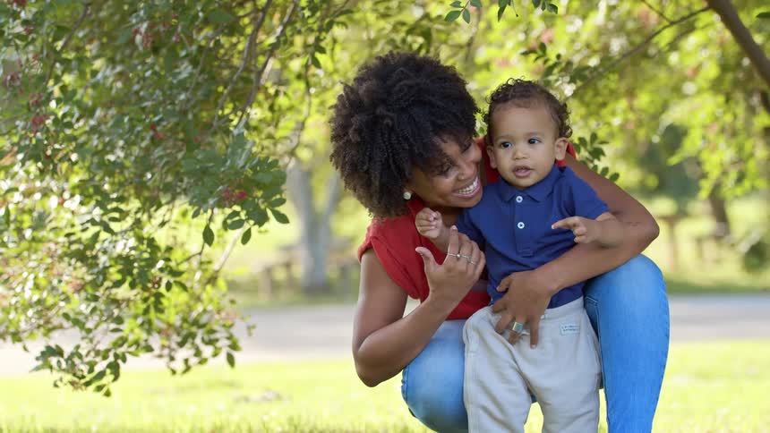 A mother holds her toddler son on the lawn as he tries to grab a tree branch but she gently directs his attention forward with a smile on her face around midday