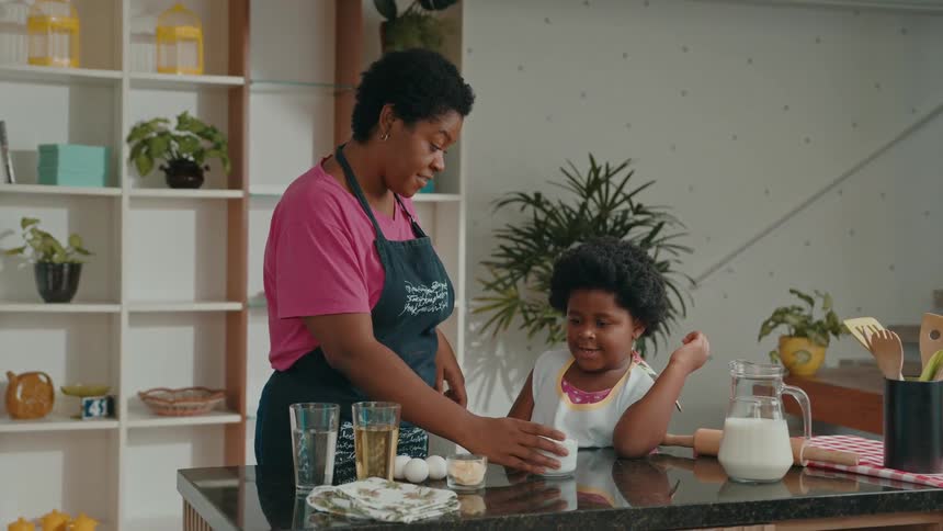 A mother and her young daughter stand in a kitchen preparing to cook with a glass pitcher of milk various ingredients and utensils arranged on the counter
