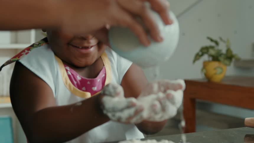 A child joyfully plays with flour while an adult pours more onto their hands in a kitchen