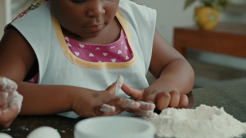Child with dark skin in a white and yellow apron over a pink shirt cleans flour from her fingers beside a small pile on the counter