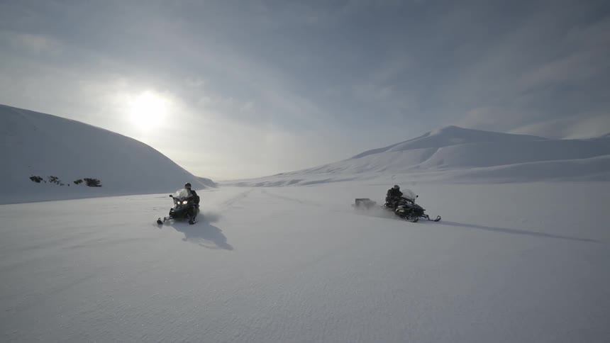 Two snowmobilers drive across a frozen landscape