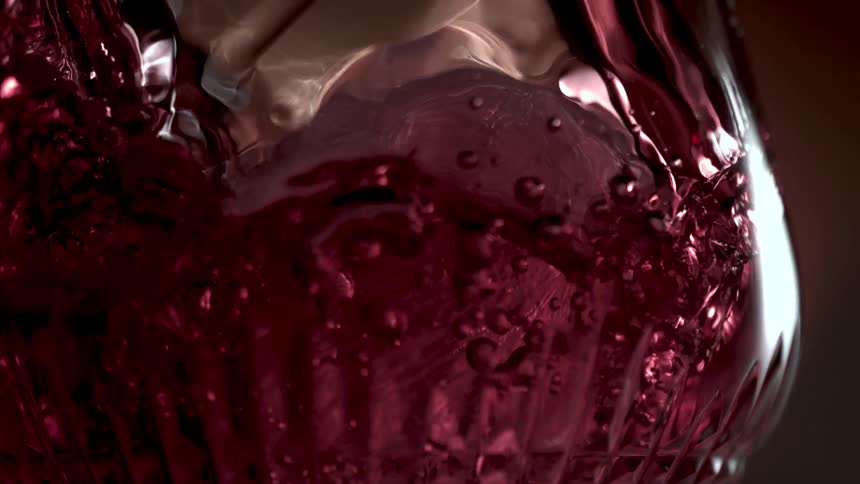 In an extreme closeup slowmotion shot a red beverage is poured over ice cubes in an ornate glass creating a turbulent swirl