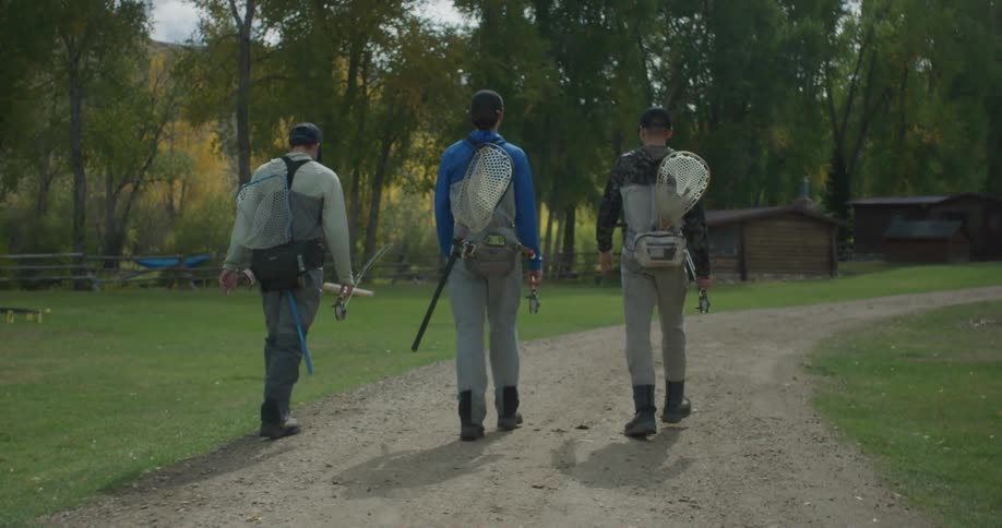 Three people walk down a dirt path carrying fishing gear in a wooded outdoor setting