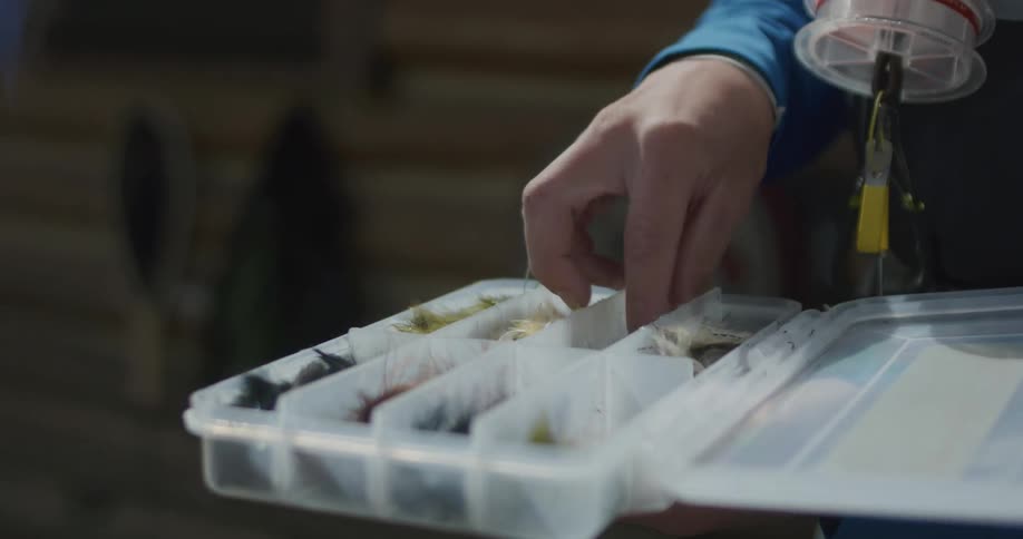 A person selects a fishing fly from a plastic tackle box with multiple compartments each holding a variety of colorful flies for fly fishing