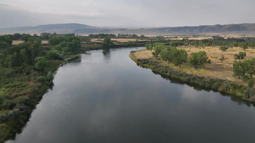 A calm river winds through a grassy landscape with trees and distant hills