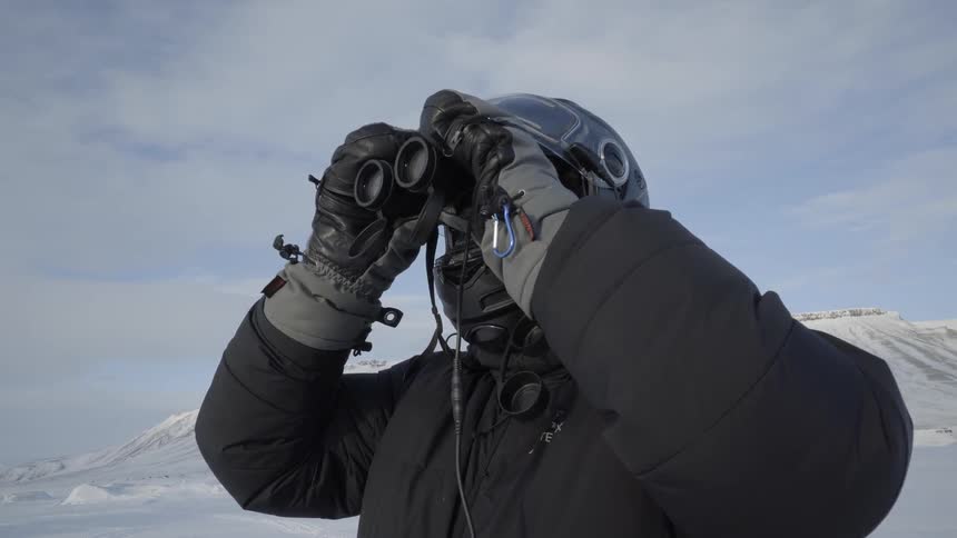 A man wearing a helmet and winter gear looks through binoculars