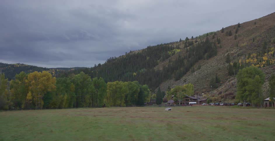 Grassy valley with autumn trees mountains and cabins under cloudy sky