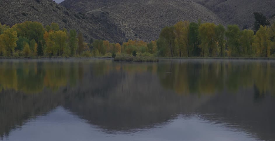 A calm lake reflects autumn colored trees and surrounding hills under an overcast sky