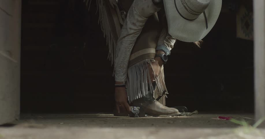 A cowboy adjusts his spur while crouching in western gear